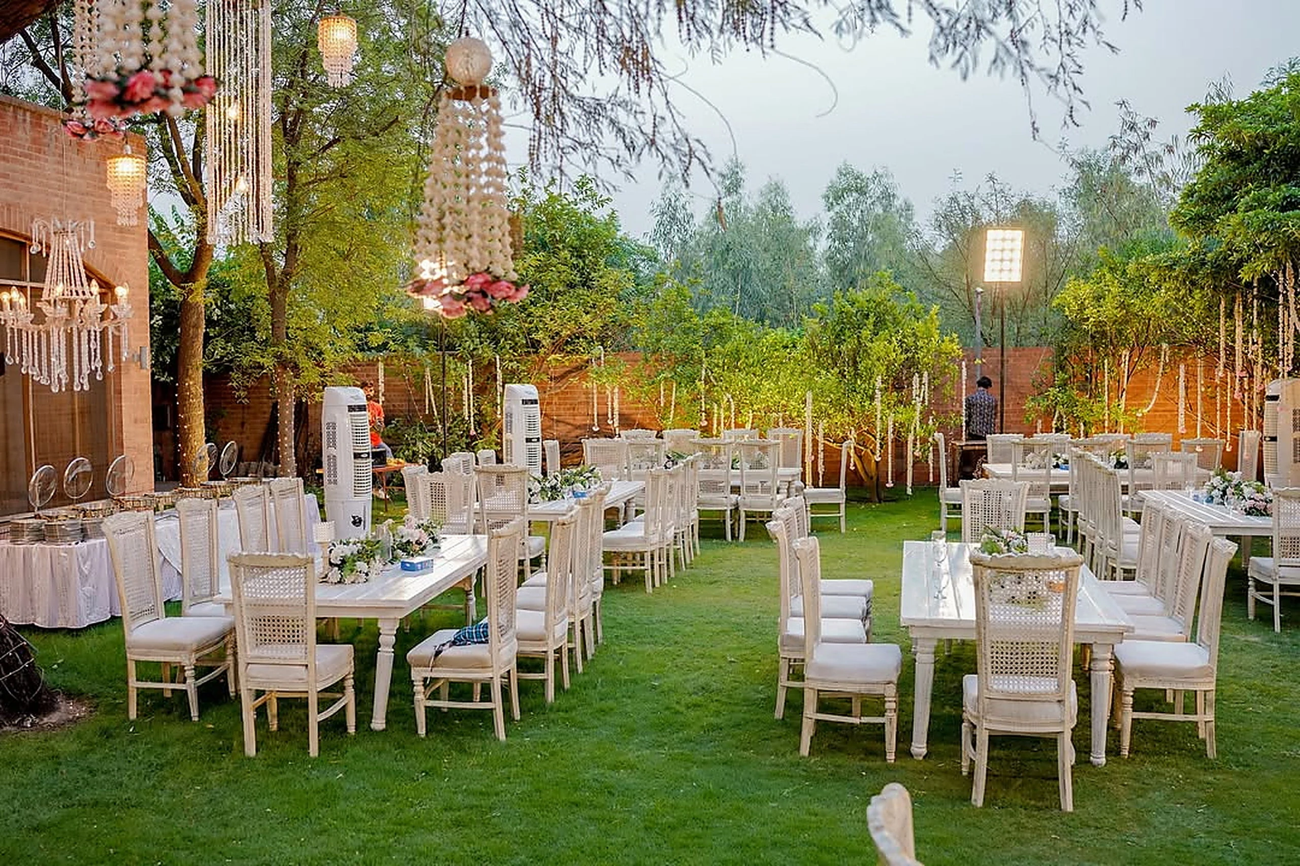 Long banquet tables dressed in white on a garden lawn beneath lantern posts